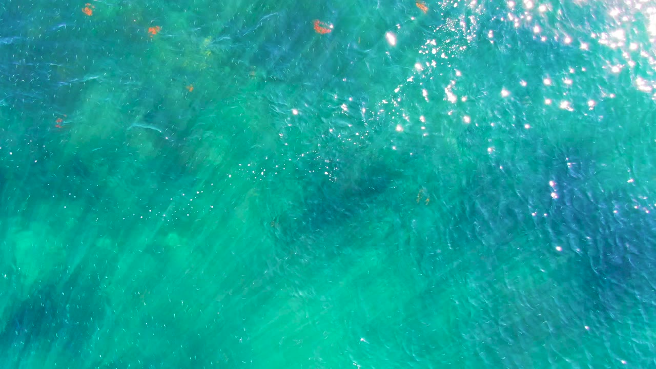 Blue-green ocean water in La Bocana fills the frame from above, with scattered small orange objects, shimmering highlights, and no visible people.