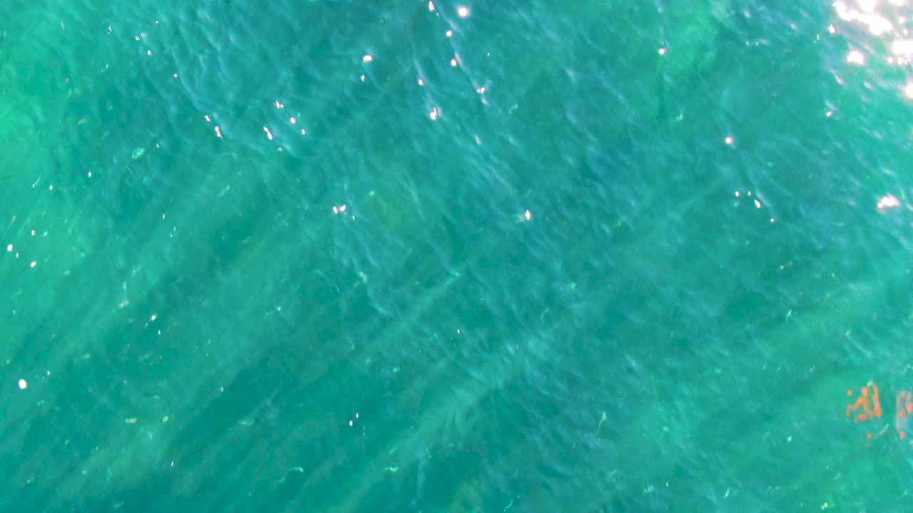 Top-down view of clear turquoise water in La Bocana, Baja California Sur, with ripples, bright sun reflections, faint seabed texture, and no visible people.