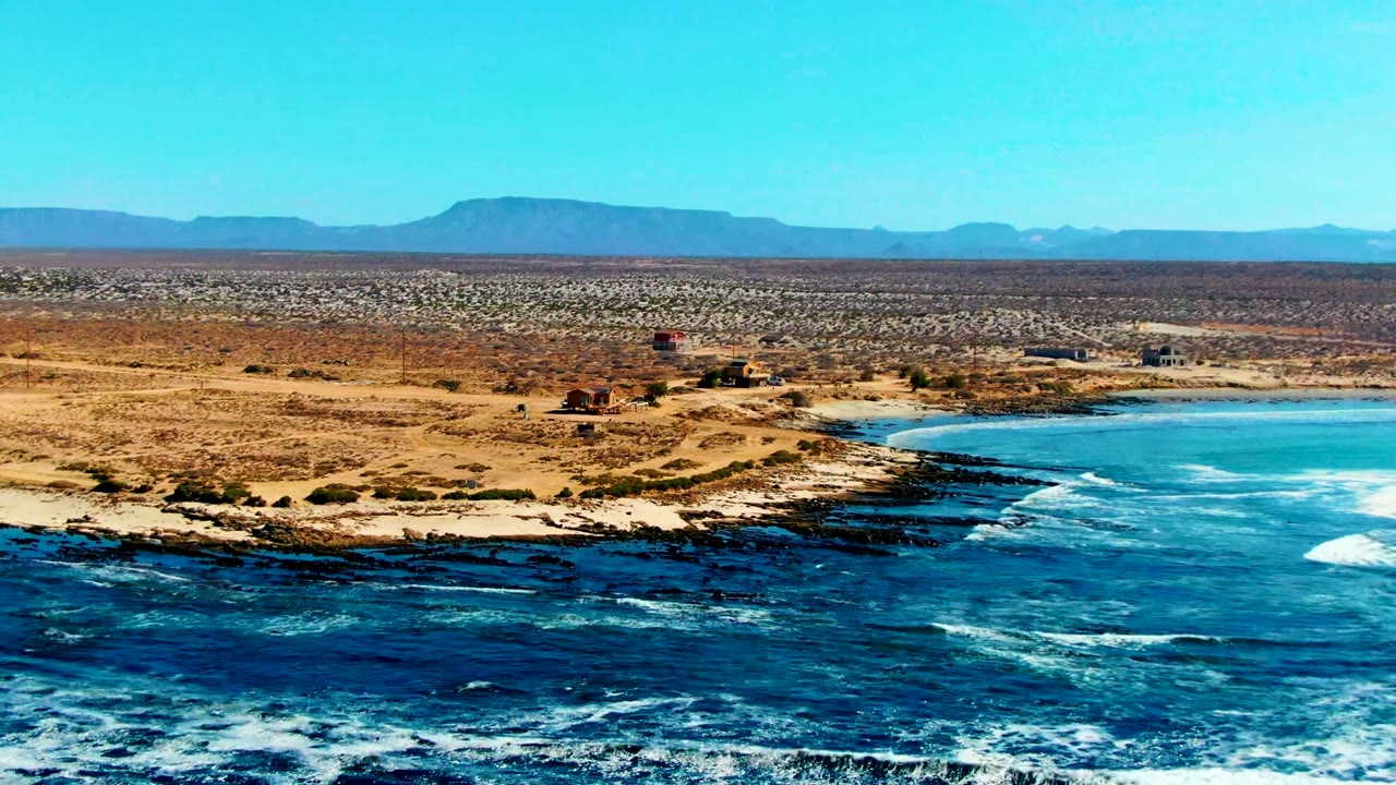 Wide aerial frame of La Bocana, Baja California Sur, with blue ocean in front, dark rock shelves along the shore, sandy desert beyond, distant mountains, and no visible people.
