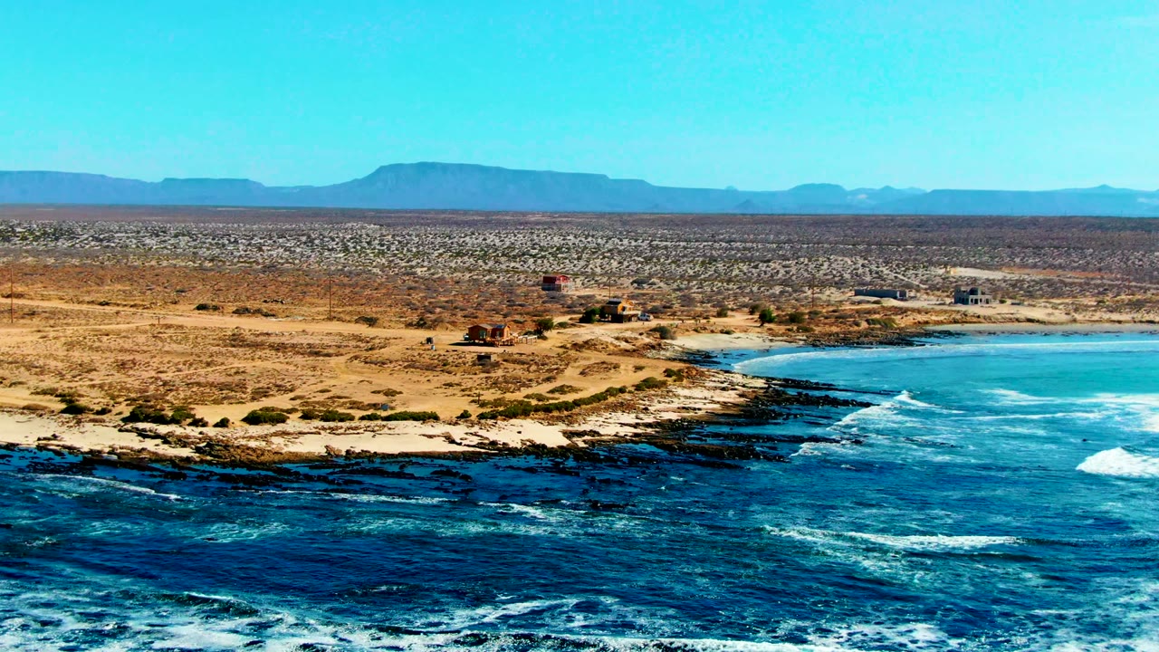 Blue surf curls toward dark rocks beside a sandy coastal desert.