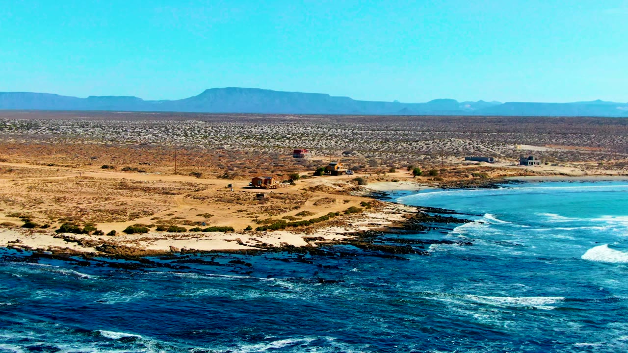 La Bocana coastline from above, showing turquoise water and white wave lines at right, black rock ledges at the shore, dry desert land with small structures, and no visible people.