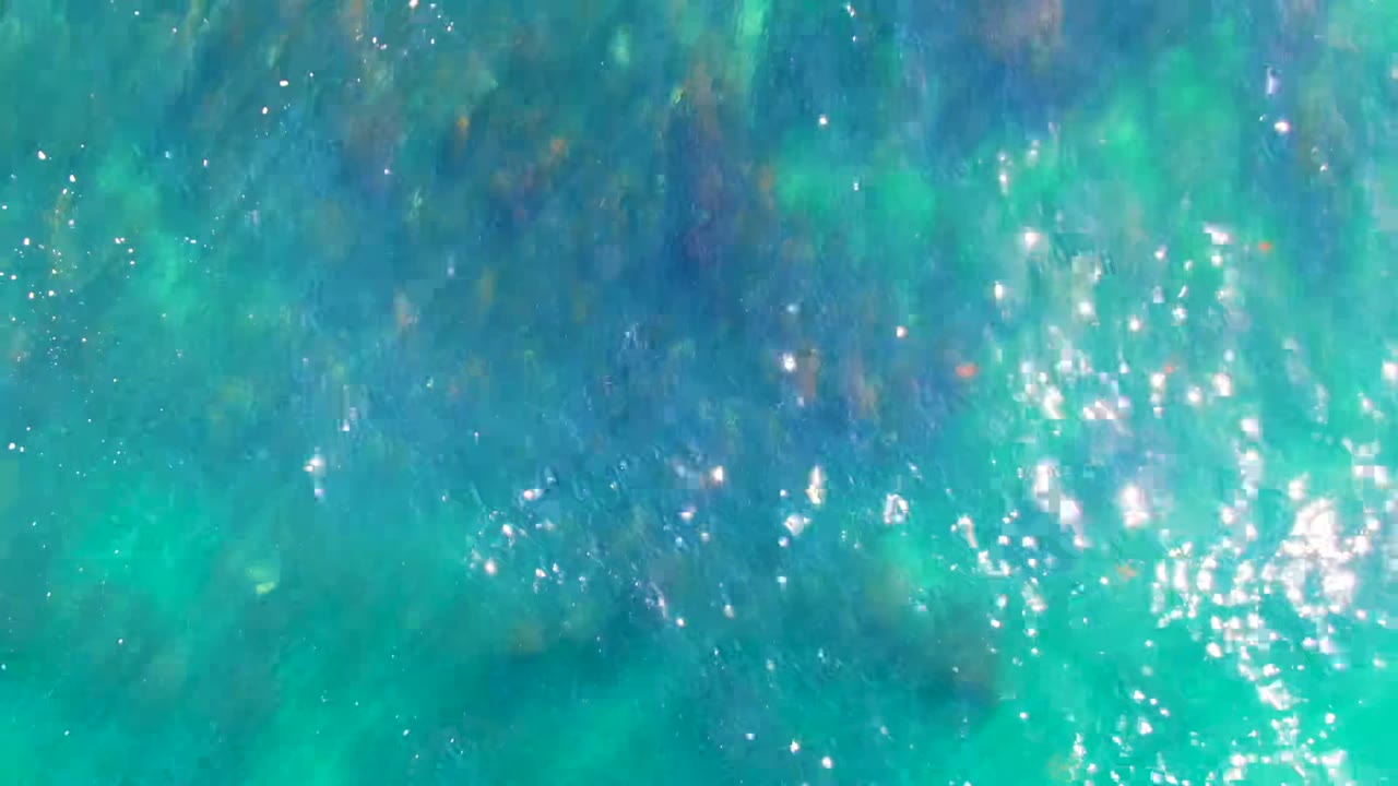 Overhead view of turquoise ocean water in La Bocana, Baja California Sur, with bright sun sparkles, faint darker seabed areas, and no visible people.