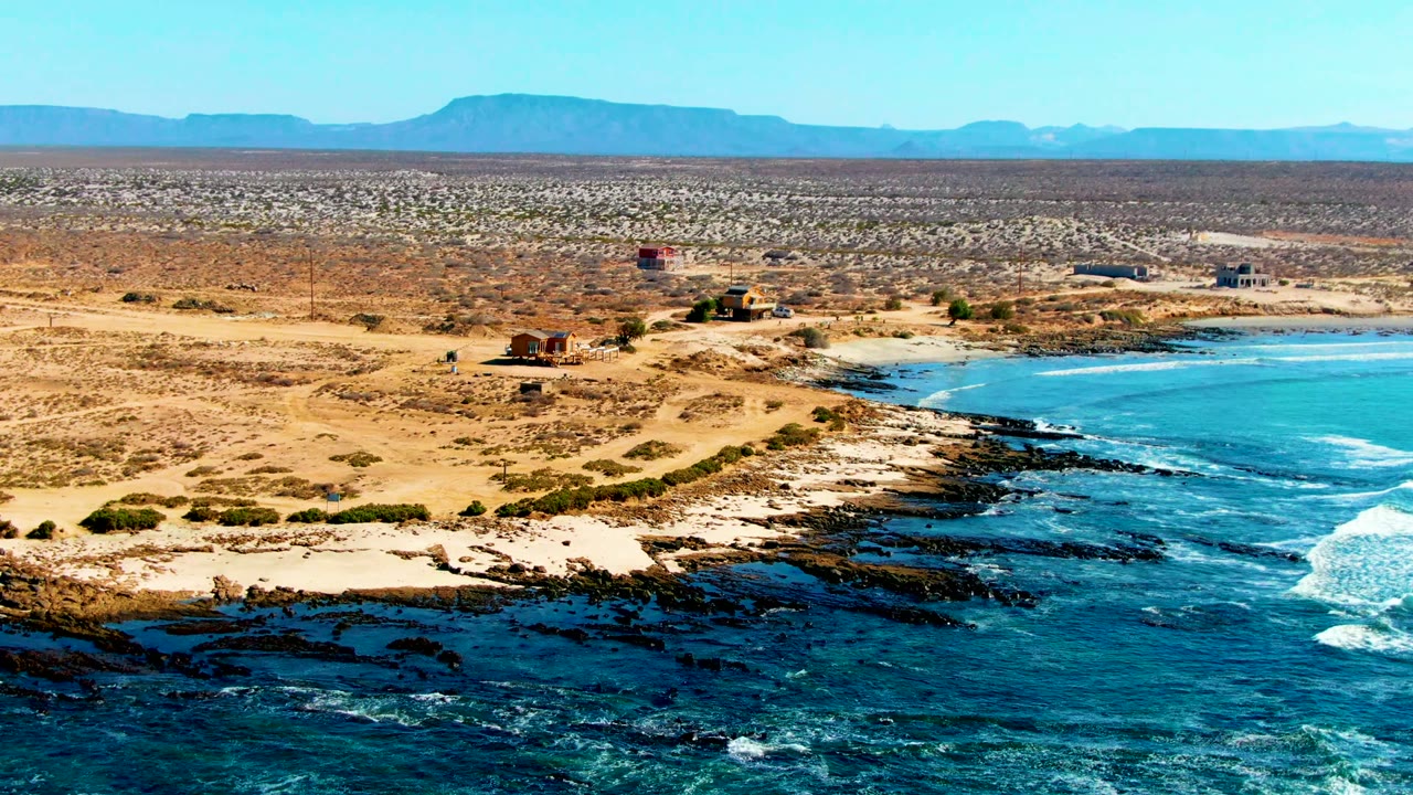 Aerial view over La Bocana, Baja California Sur, with dark rocks and blue surf in the foreground, sandy desert and small structures behind, distant mountains, and no visible people.