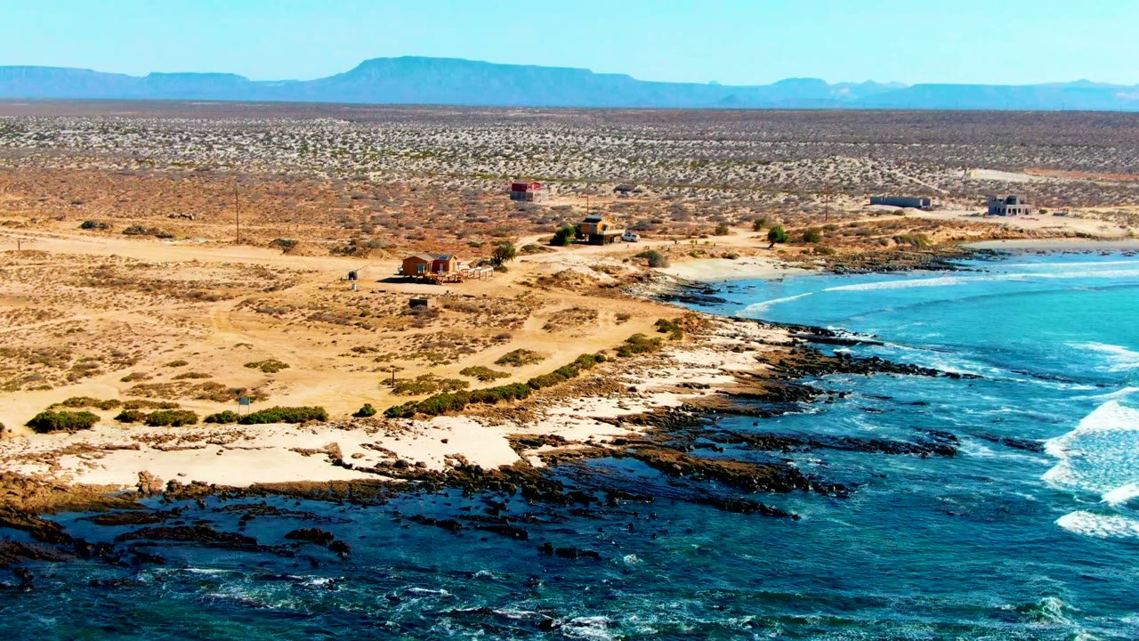 Drone view of turquoise water, rock shelves, sand, desert roads, and distant mesas.