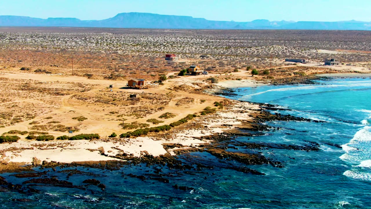 Aerial frame over La Bocana with the blue bay filling the right side, black shoreline rocks below, sandy tracks and small structures across the desert, and no visible people.