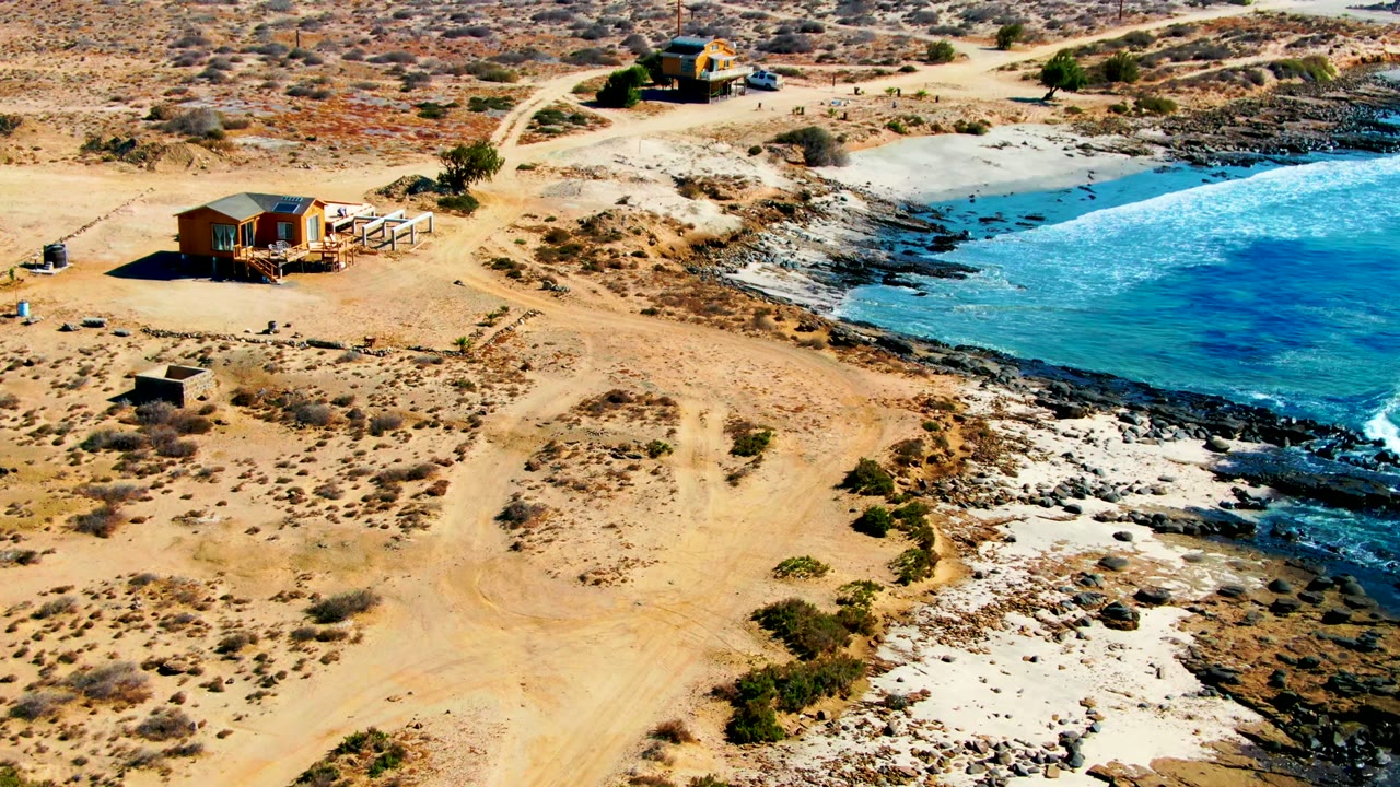 Aerial frame over La Bocana, Baja California Sur, showing a sandy desert road, a small shoreline house, rocky beach, blue water, and no visible people.