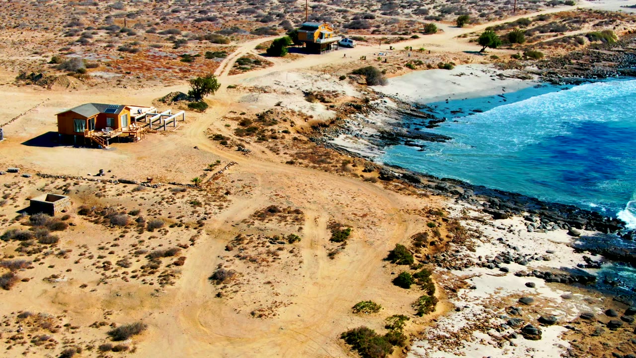 Drone view of a sandy road curving past a small coastal house and rocky beach.