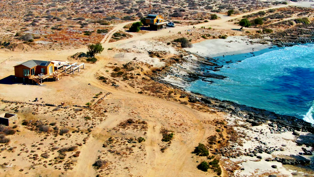 La Bocana aerial view with a cabin near the left edge, branching dirt tracks across arid ground, pale rocky shoreline, turquoise water, and no visible people.