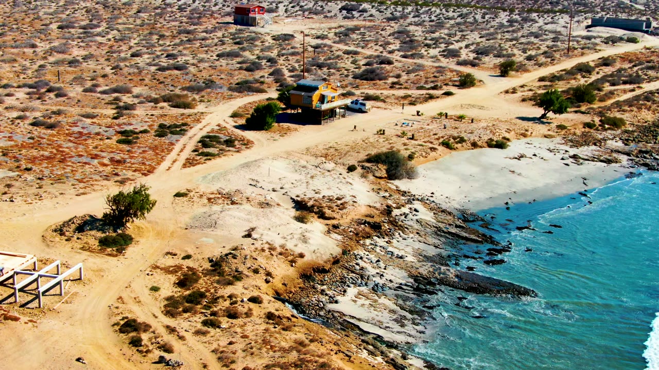 Aerial frame from La Bocana, Baja California Sur, Mexico, showing intersecting dirt roads, a small yellow building, a blue car, scrub desert, and turquoise water along rocks with no visible people.