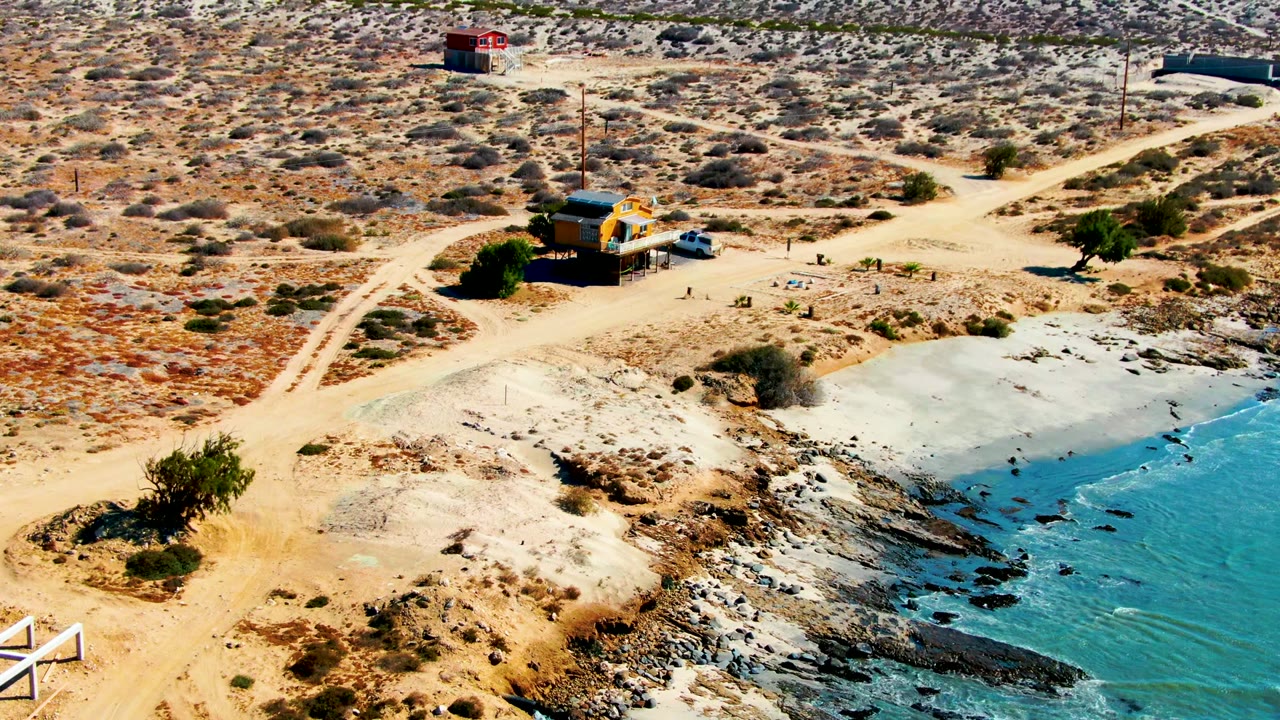 High aerial view of a yellow coastal building near dirt tracks and turquoise water.