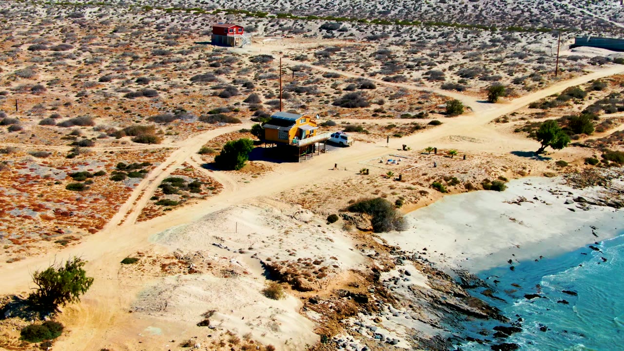La Bocana aerial frame with more desert road pattern visible behind the small yellow building, a red structure in the background, and blue water washing against the rocky shore with no visible people.