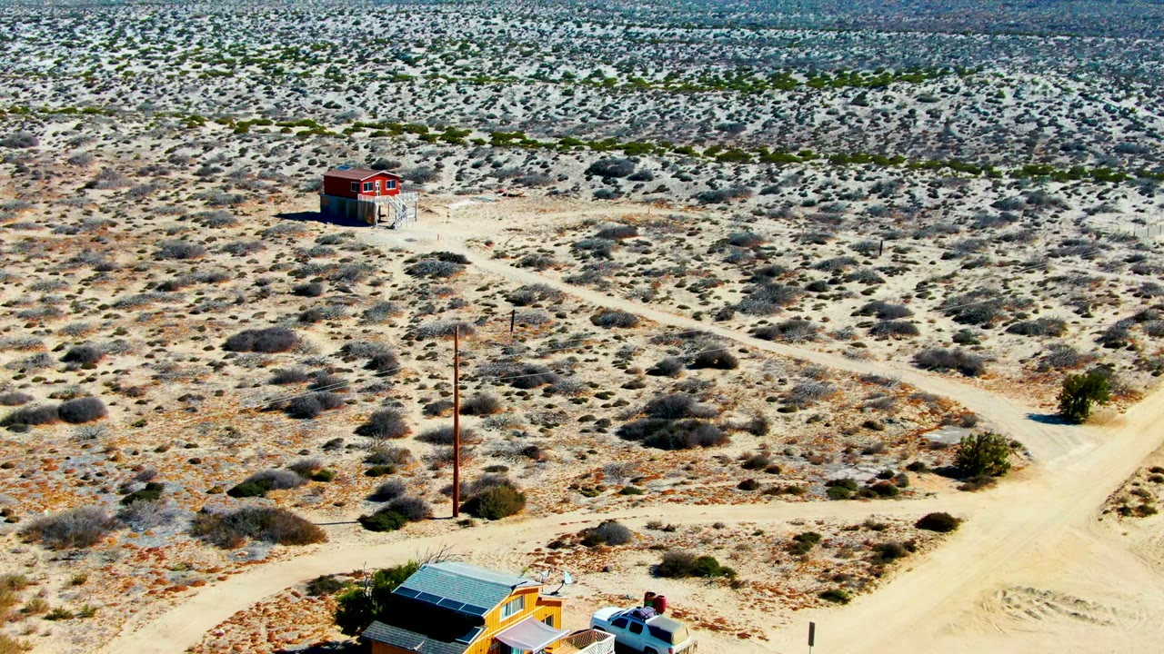 Aerial view over La Bocana desert scrub with a small red building in the upper left, intersecting dirt roads, a utility pole, and no visible people.