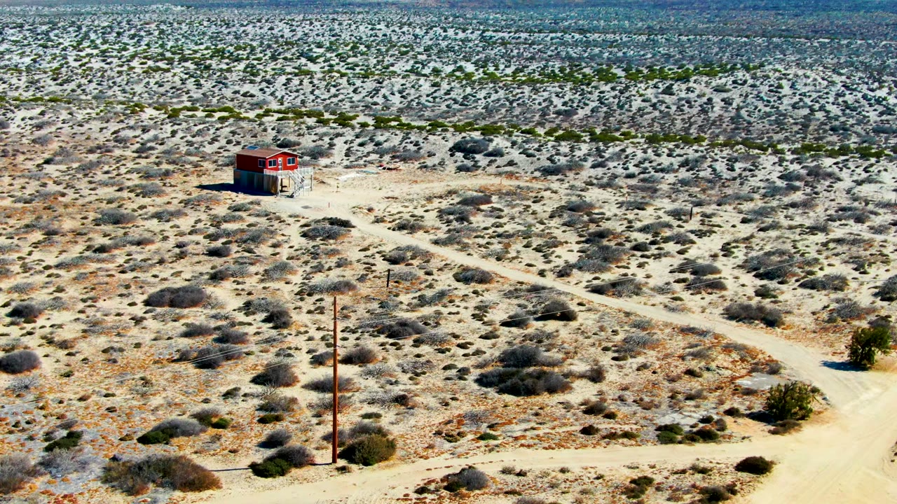 La Bocana desert aerial with the red building near the upper center, pale vehicle tracks crossing the scrub, a foreground utility pole, and no visible people.
