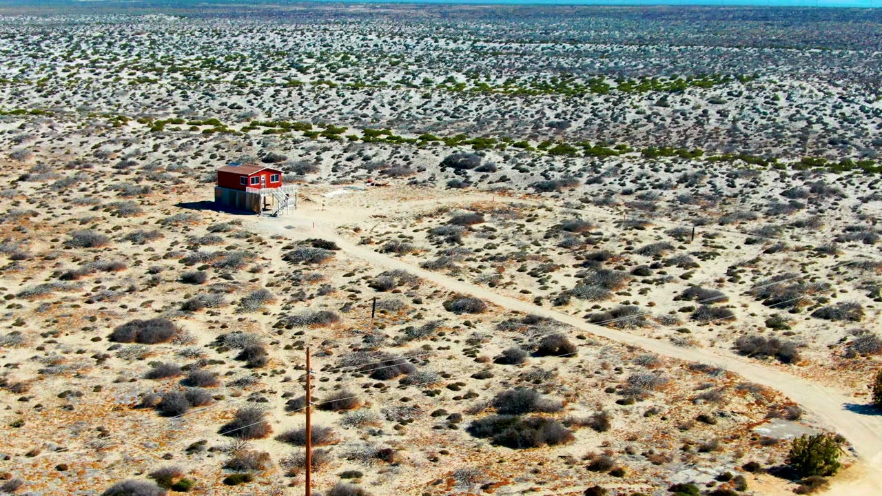 Wide aerial over La Bocana, Baja California Sur, showing a red building to the upper left of a dirt road junction, scattered shrubs, distant mountains, and no visible people.