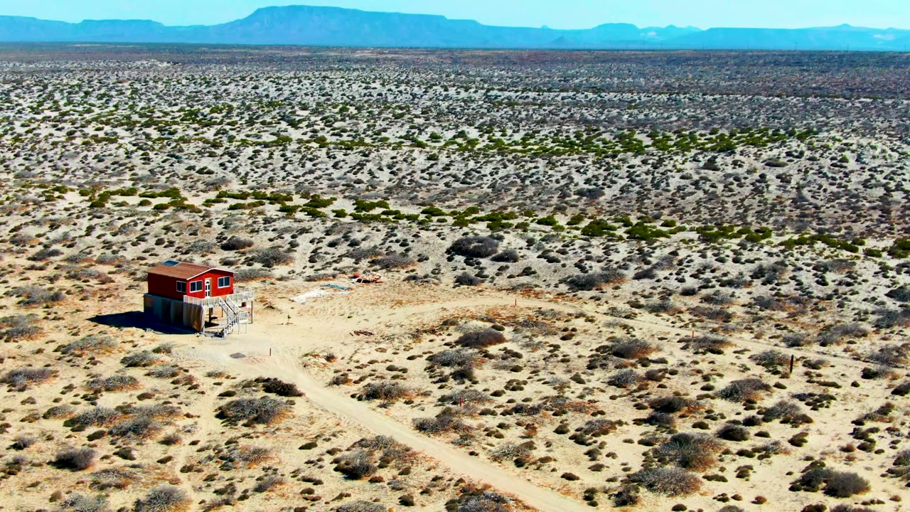 At 0:00, a small red structure sits low in the left side of a wide La Bocana desert aerial view, with dirt tracks, scattered shrubs, distant mountains, clear sky, and no visible people.