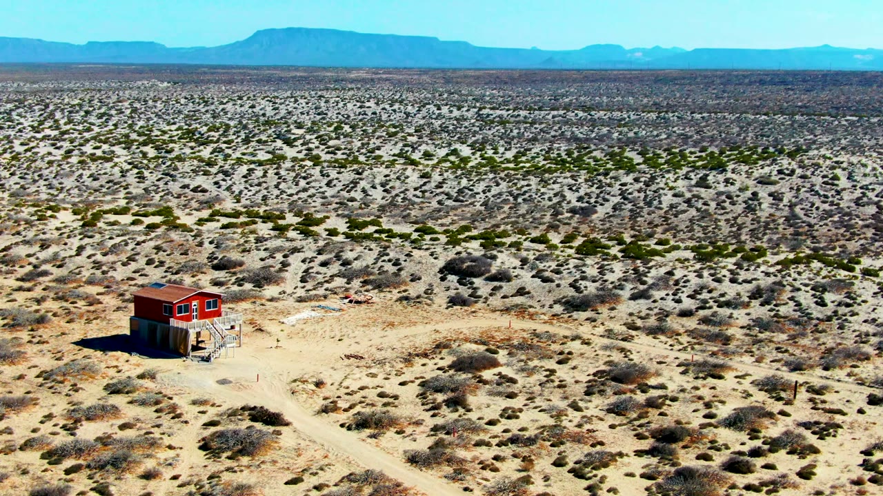At 0:02, the red structure sits slightly right of center in a wide La Bocana aerial frame, surrounded by dry brush, pale tracks, distant blue mountains, and no visible people.