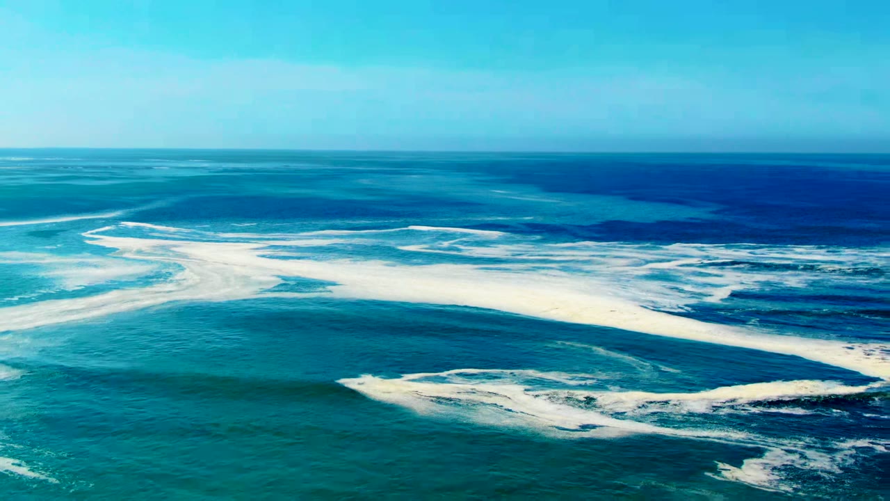 Aerial view over the ocean at La Bocana, Baja California Sur, with turquoise water, white foam streaks, a straight horizon, and no visible people.
