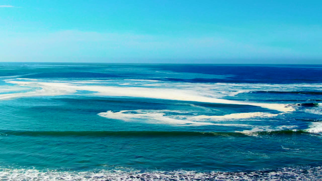 Foamy ocean waves roll toward shore beneath a clear blue sky.
