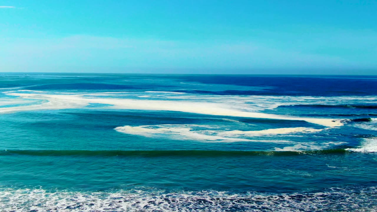 At La Bocana, Baja California Sur, white foam arcs across the middle of the turquoise surf while a darker blue band of open ocean sits near the horizon, with no visible people.