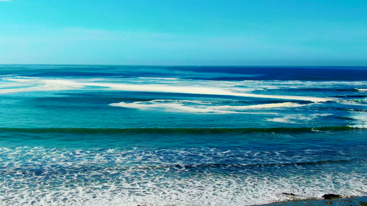 Wide elevated view of turquoise and deep blue ocean bands at La Bocana, Baja California Sur, with white waves rolling toward shore and no visible people.