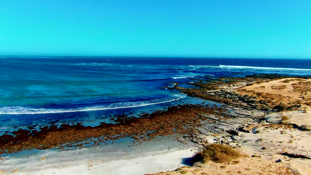 Aerial view over La Bocana, Baja California Sur, with turquoise water, dark rocky reef, pale sand at right, a clear horizon, and no visible people.