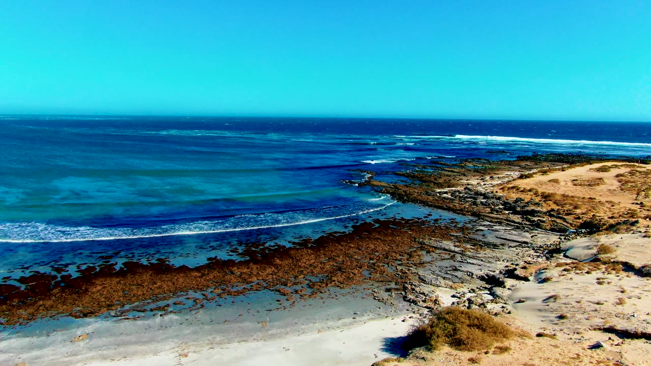Blue ocean and white surf crossing a rocky coastal shelf beside sand.