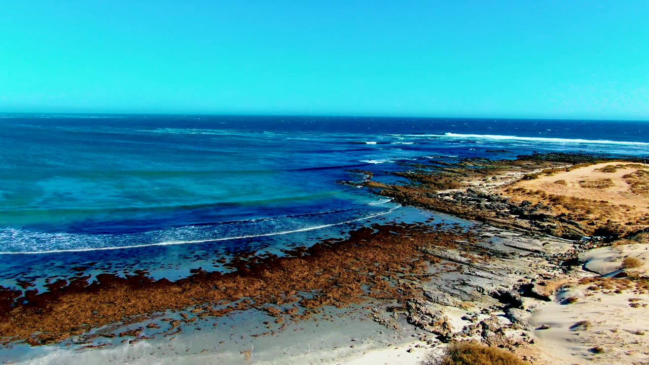 High coastal view at La Bocana showing turquoise shallows on the left, a dark reef band through the middle, sandy shore at right, and no visible people.