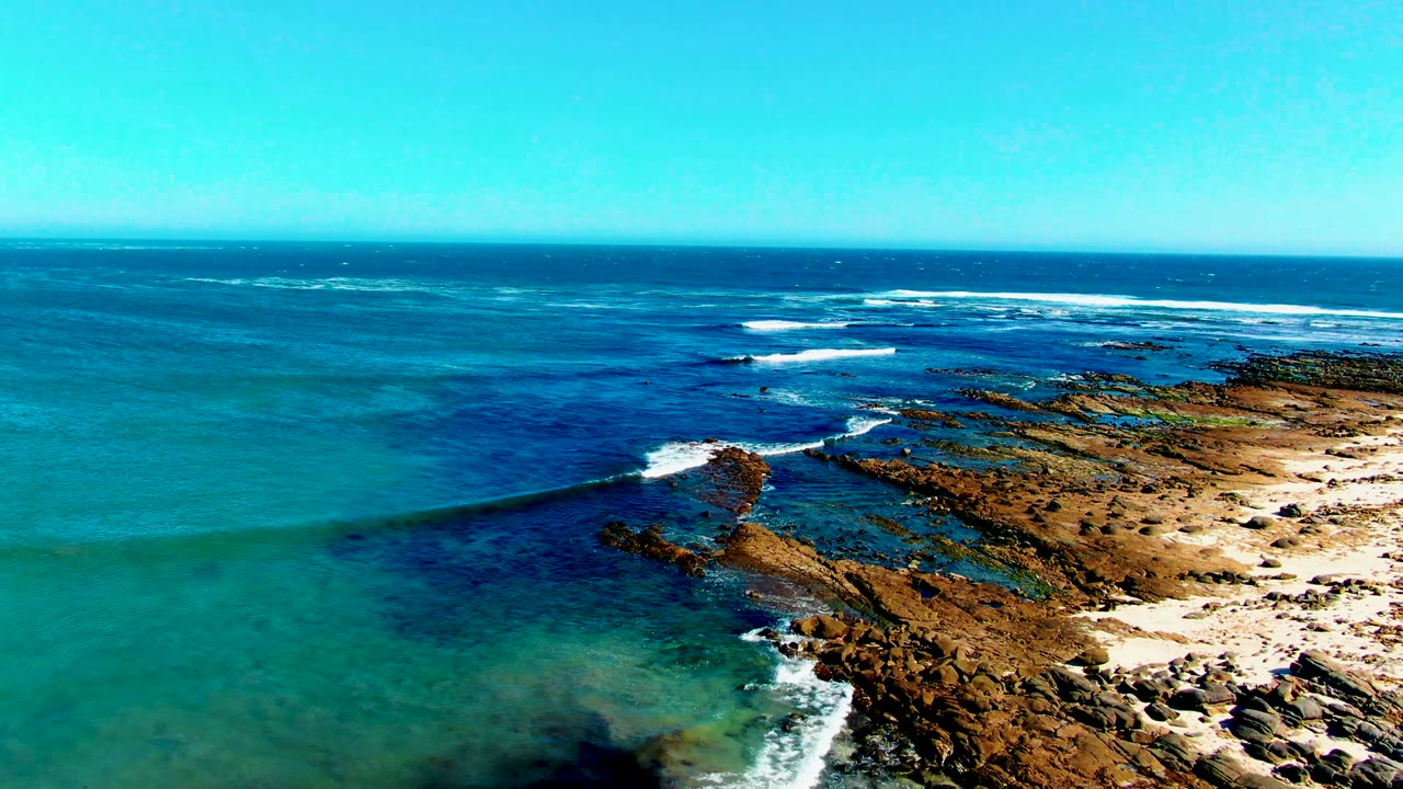 Aerial view over La Bocana, Baja California Sur, showing deep blue ocean, turquoise shallows, rocky shore, sandy patches, and no visible people.