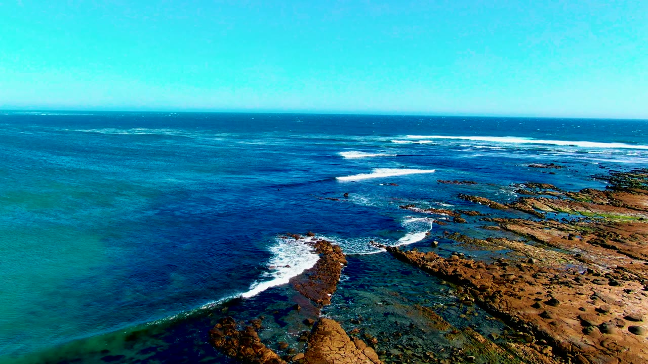 Aerial view at La Bocana showing white surf curling around a narrow rock formation, sandy shoreline to the right, blue ocean beyond, and no visible people.