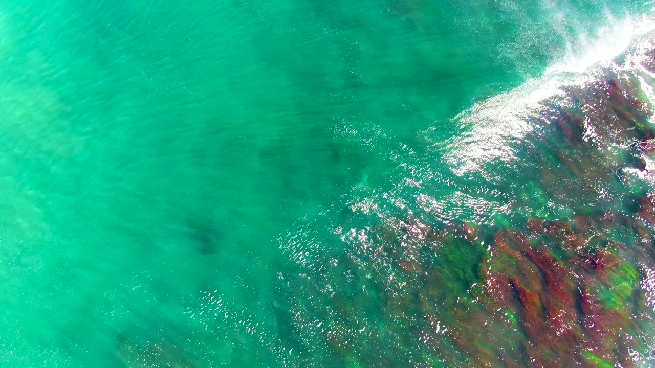 Overhead view of turquoise water at La Bocana, Mexico, where white surf enters from the upper right over dark submerged rocks, with no visible people.