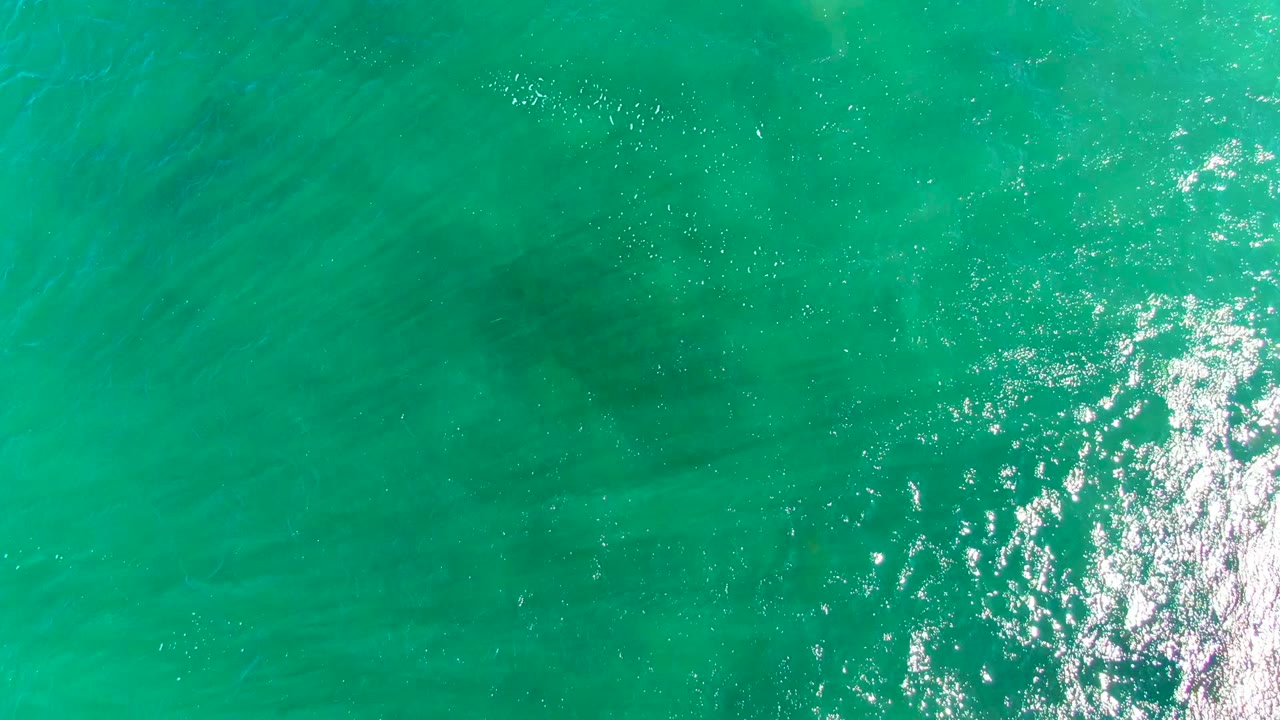 Overhead view in La Bocana with clear turquoise water, fine diagonal ripples, a concentrated bright reflection on the right side, and no visible people.