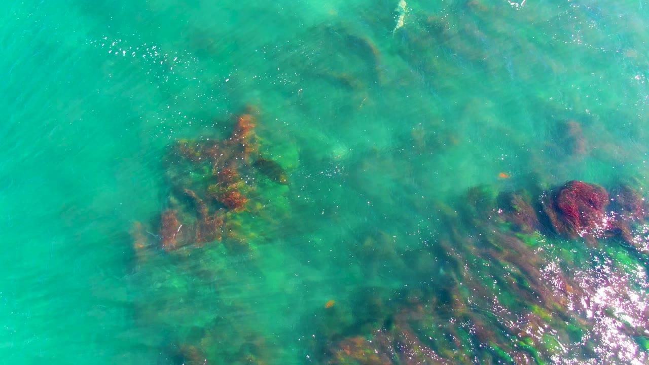 Overhead view of turquoise water in La Bocana, Baja California Sur, with dark and reddish submerged rocks visible near the lower right and no visible people.