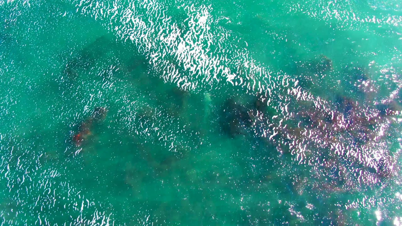 Overhead view in La Bocana showing turquoise water, fine white ripple highlights, and darker underwater rock forms crossing the upper and center areas, no visible people.