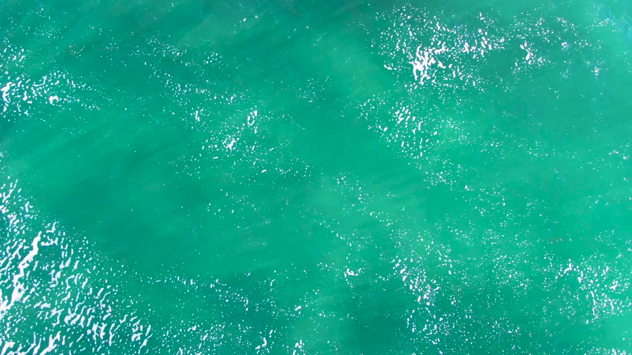 Overhead turquoise ocean at La Bocana, Baja California Sur, shows brighter clusters of reflected sunlight across the central water surface, no visible people.
