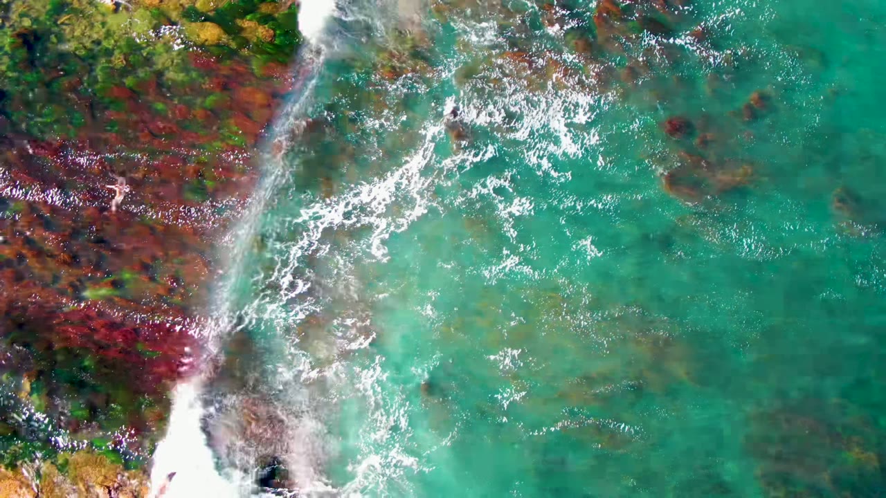 Overhead view at La Bocana, Baja California Sur, with turquoise water, white foam, and red-brown rocks along the left edge, with a tiny swimmer shape near the rocky shallows.