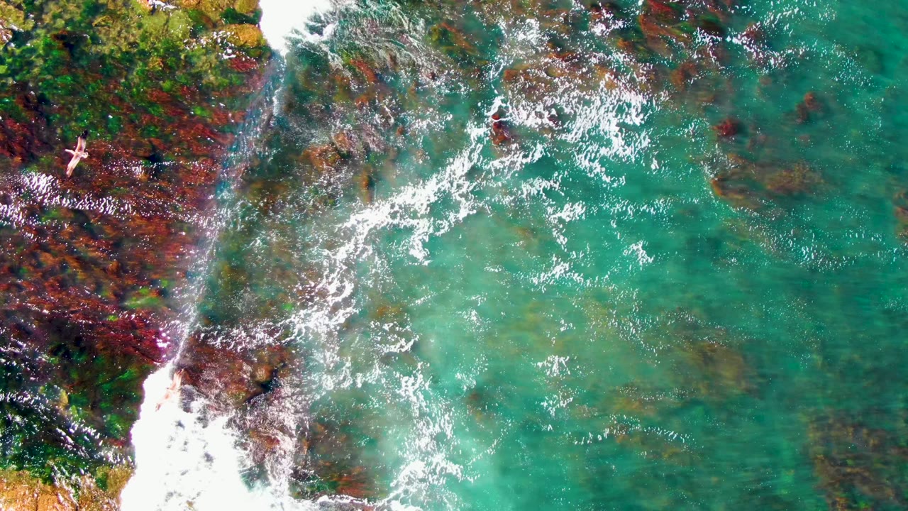 Aerial frame over La Bocana shallow coastal water where a bright foam band crosses red-brown rock and green-blue water, with the swimmer still very small near the left rocky area.