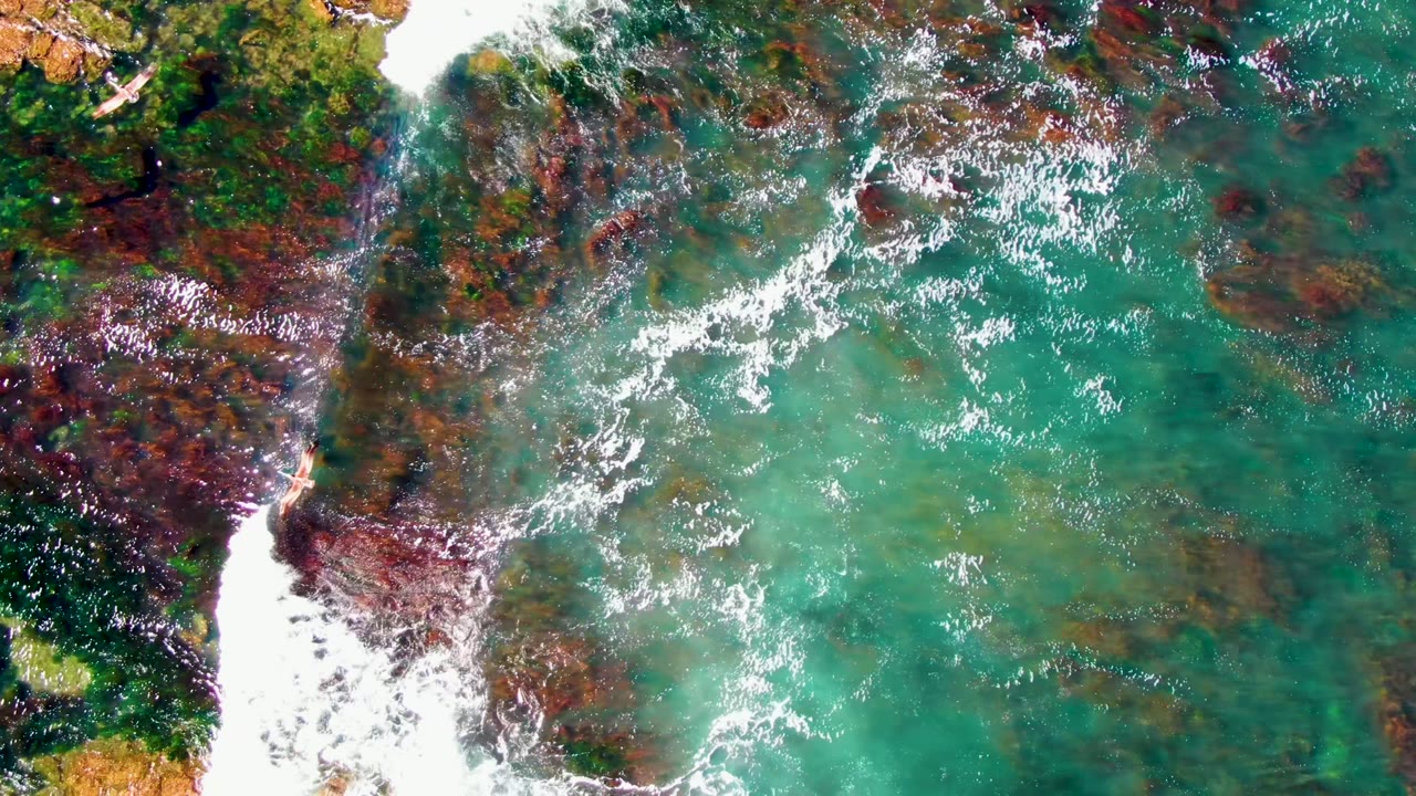 La Bocana shoreline from above with a broad white surf patch at left, mottled red-brown rock under clear turquoise water, and a small swimmer visible near the rock edge.