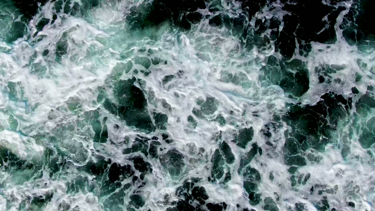 Top-down frame of white surf foam spreading across dark green ocean water and submerged rocks at La Bocana, Baja California Sur, with no visible people.