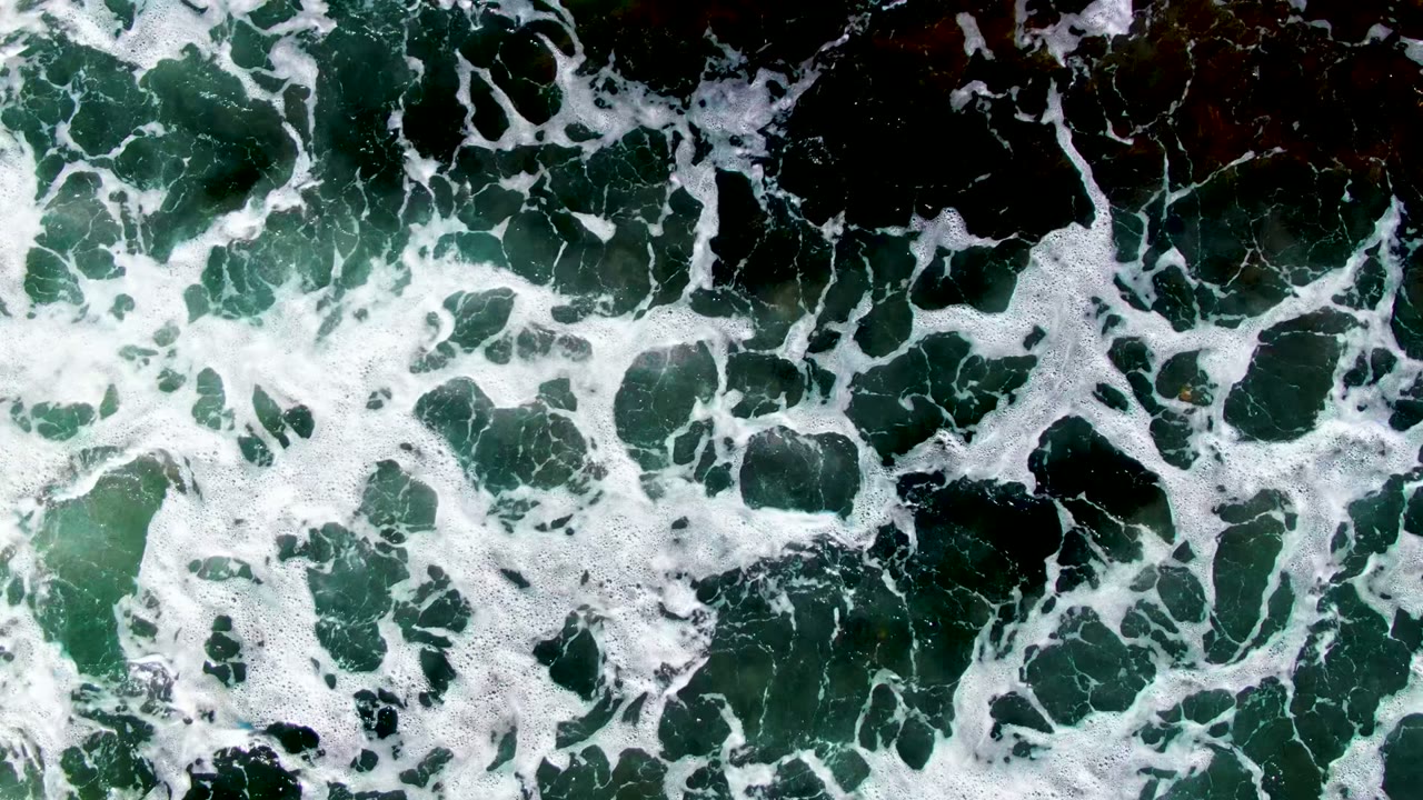 Top-down surf frame at La Bocana, Baja California Sur, where white foam sheets and channels cover dark green coastal water, with no visible people.