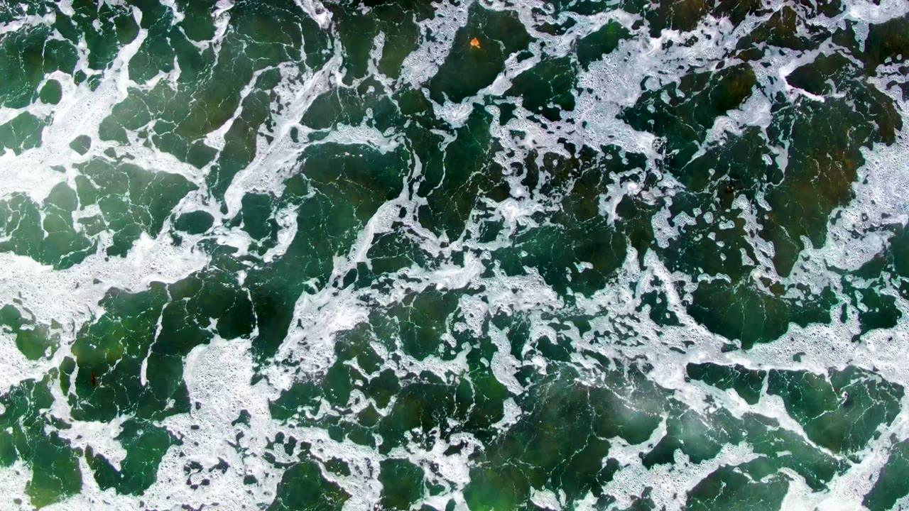 Overhead view in La Bocana, Baja California Sur, Mexico, with white foam streaks crossing green shallow ocean water and submerged seaweed, no visible people.