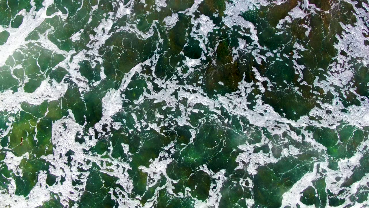 Aerial frame from La Bocana showing denser white surf foam over dark green water with brown seaweed visible beneath, no visible people.