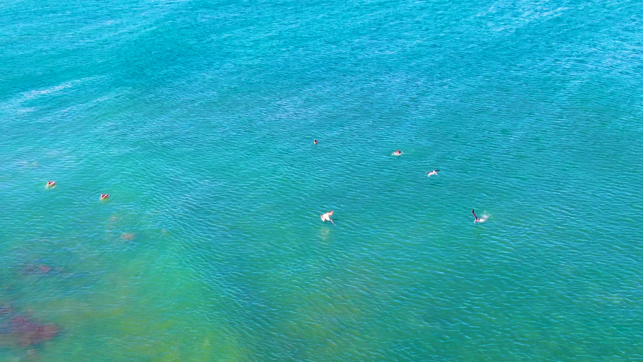 Overhead La Bocana water view with swimmers spaced across the upper and middle frame, textured ripples, and darker seabed patches in the lower water.