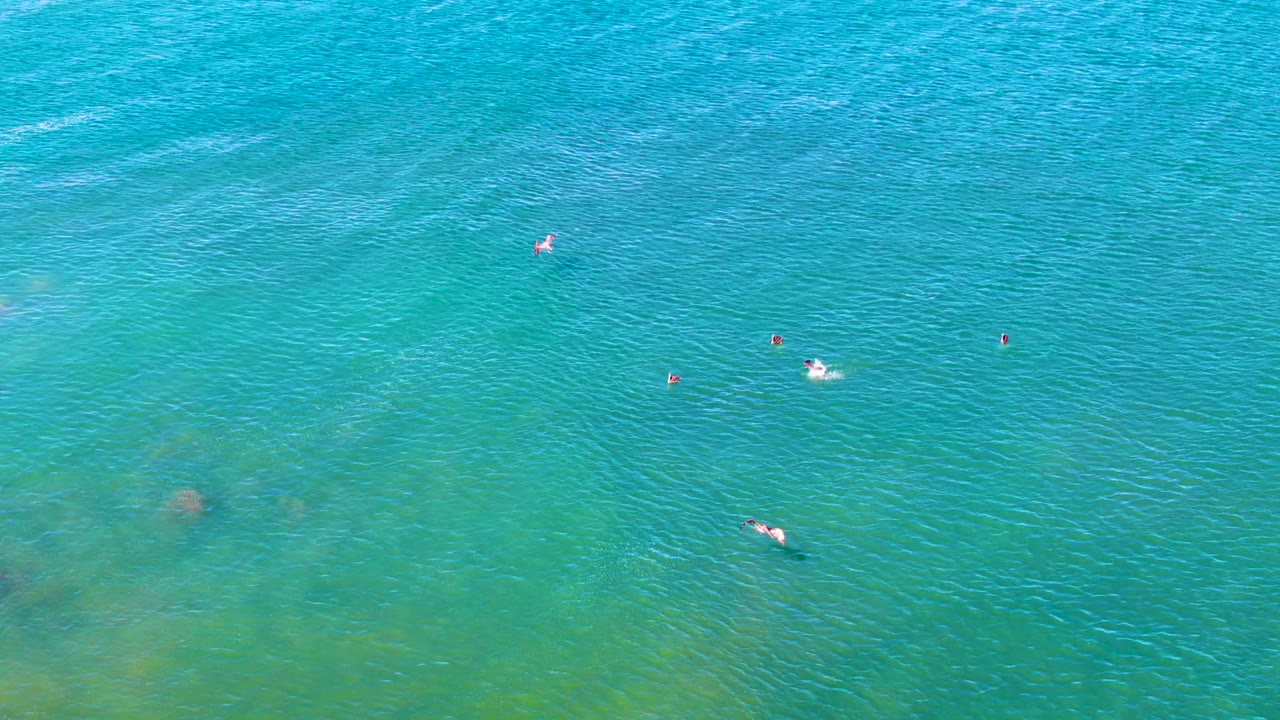 Overhead La Bocana water frame with scattered swimmers, visible ripples, and one bright white float or splash near the right side of the composition.