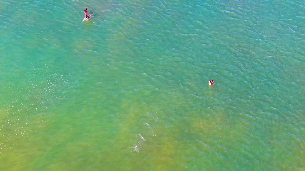 At 0:02, multiple swimmers remain visible from above in La Bocana's shallow ocean water, with a faint white splash trail near the center and green sandbar-like color shifts below the surface.