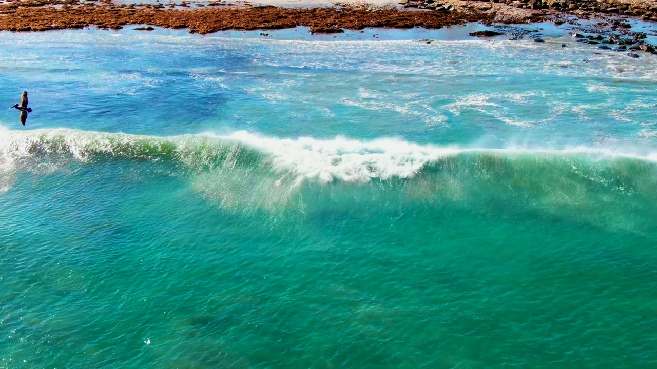 Aerial view of a wave cresting in turquoise water beside rocks and sand.