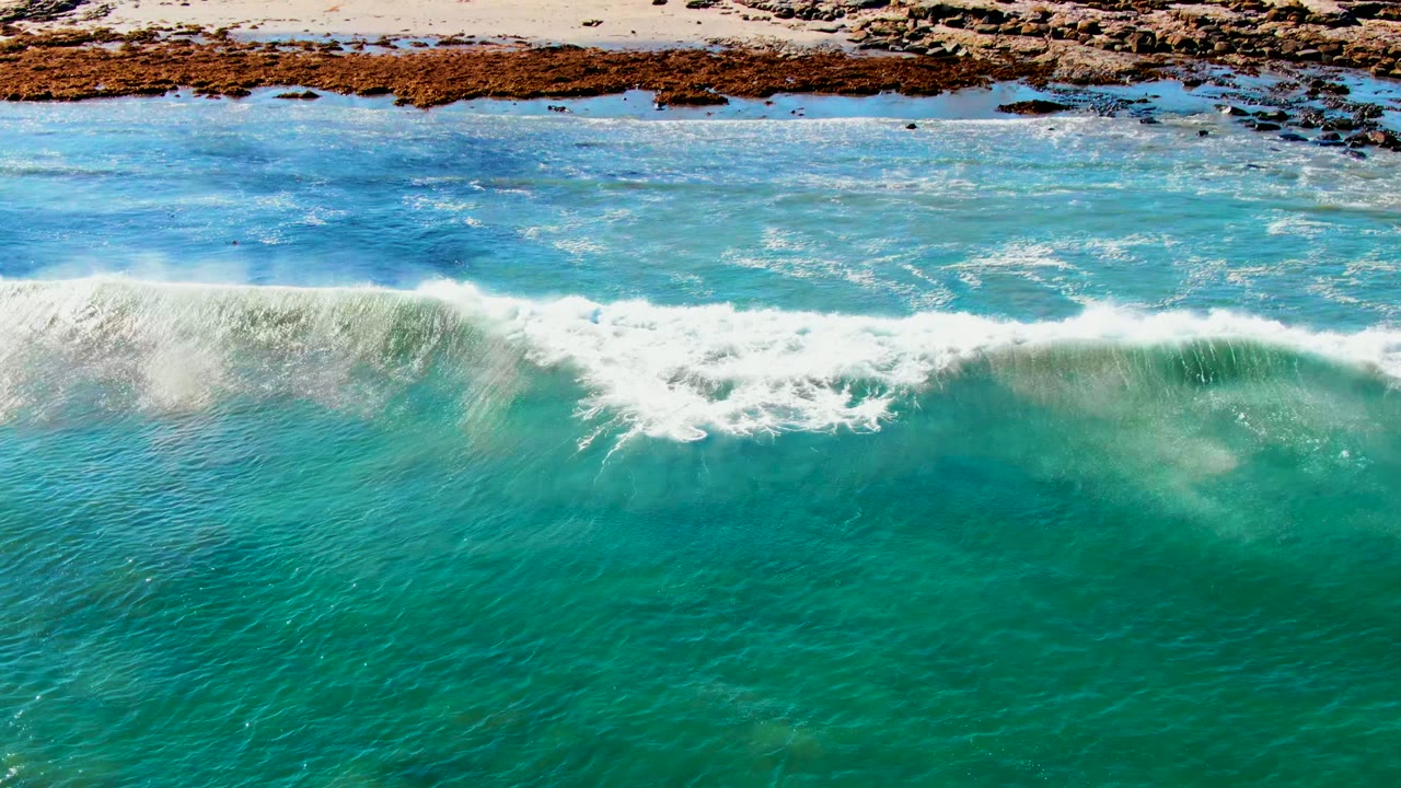 Aerial frame from La Bocana, Baja California Sur showing the wave breaking wider across turquoise water, with white spray below the rocky and sandy shoreline and no visible people.
