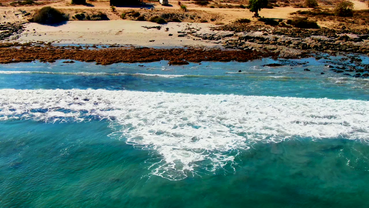 Aerial frame over La Bocana, Baja California Sur showing white surf spreading across blue-green water toward a rocky sandy shore, with dry scrubland beyond and no visible people.