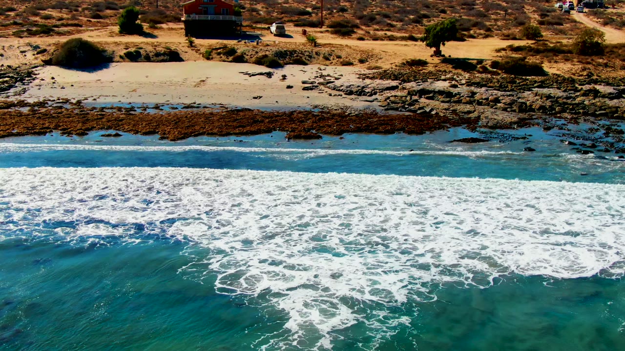 Aerial frame over La Bocana showing dense whitewater in the foreground, clear blue-green shallows near rocks, pale sand, dry brush, and small coastal buildings in the distance with no visible people.
