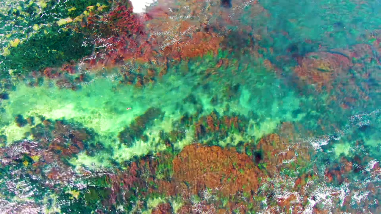 Overhead view of shallow turquoise water over red and brown rocky seabed in La Bocana, Baja California Sur, with no visible people.
