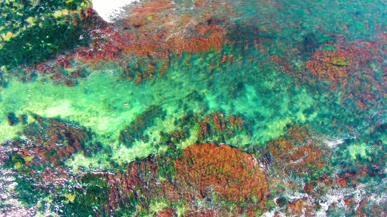 Turquoise shallows reveal reddish rock bands and darker marine texture below the surface in La Bocana, Baja California Sur, with no visible people.